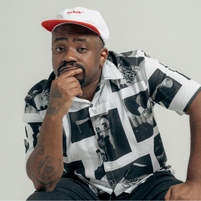 Homem negro, sentado com a mão apoiada no joelho, com boné branco e aba vermelha, veste camisa estampada preto e branca